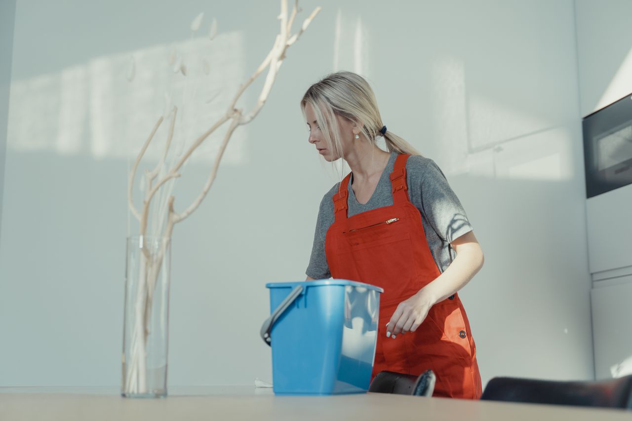 Une femme en uniforme rouge nettoie à l’intérieur avec un seau bleu, garantissant un environnement impeccable.